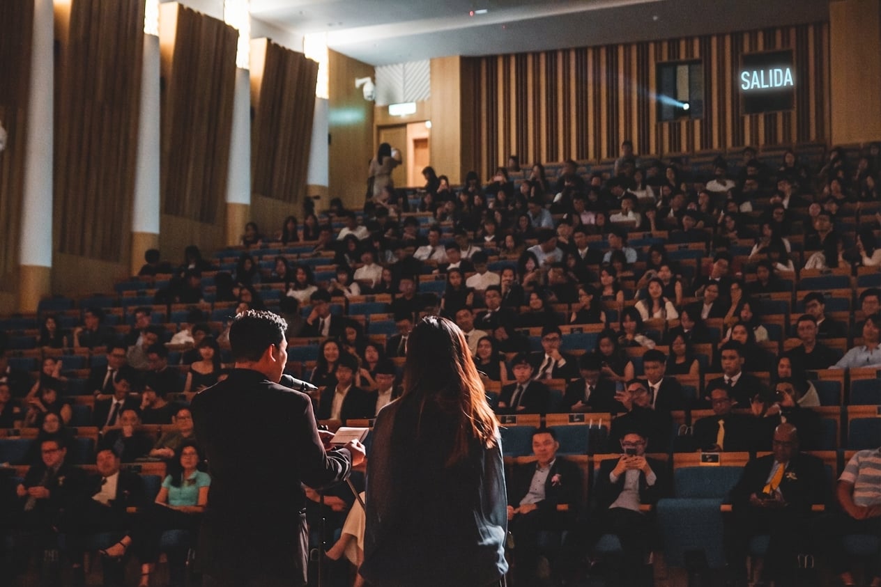 Dos personas están en el escenario hablando ante una sala de conferencias completamente llena con cientos de personas