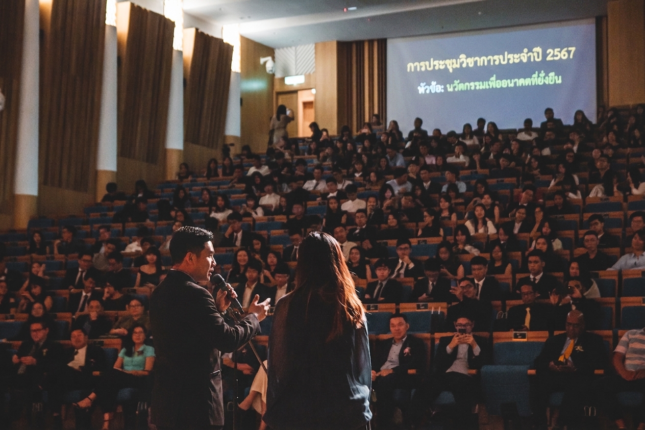 สองคนกำลังพูดบนเวทีต่อหน้าผู้ฟังในห้องบรรยายที่เต็มไปด้วยผู้คนหลายร้อยคน