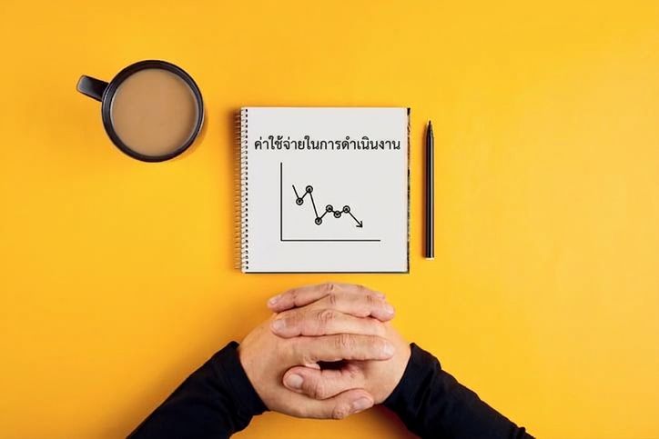 ภาพสมุดบันทึกแสดงค่าใช้จ่ายดำเนินงานที่ลดลง พร้อมแก้วกาแฟและแขนของชาย