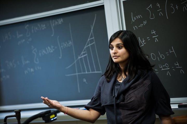 University professor giving a lecture. A blackboard full of math is behind her.