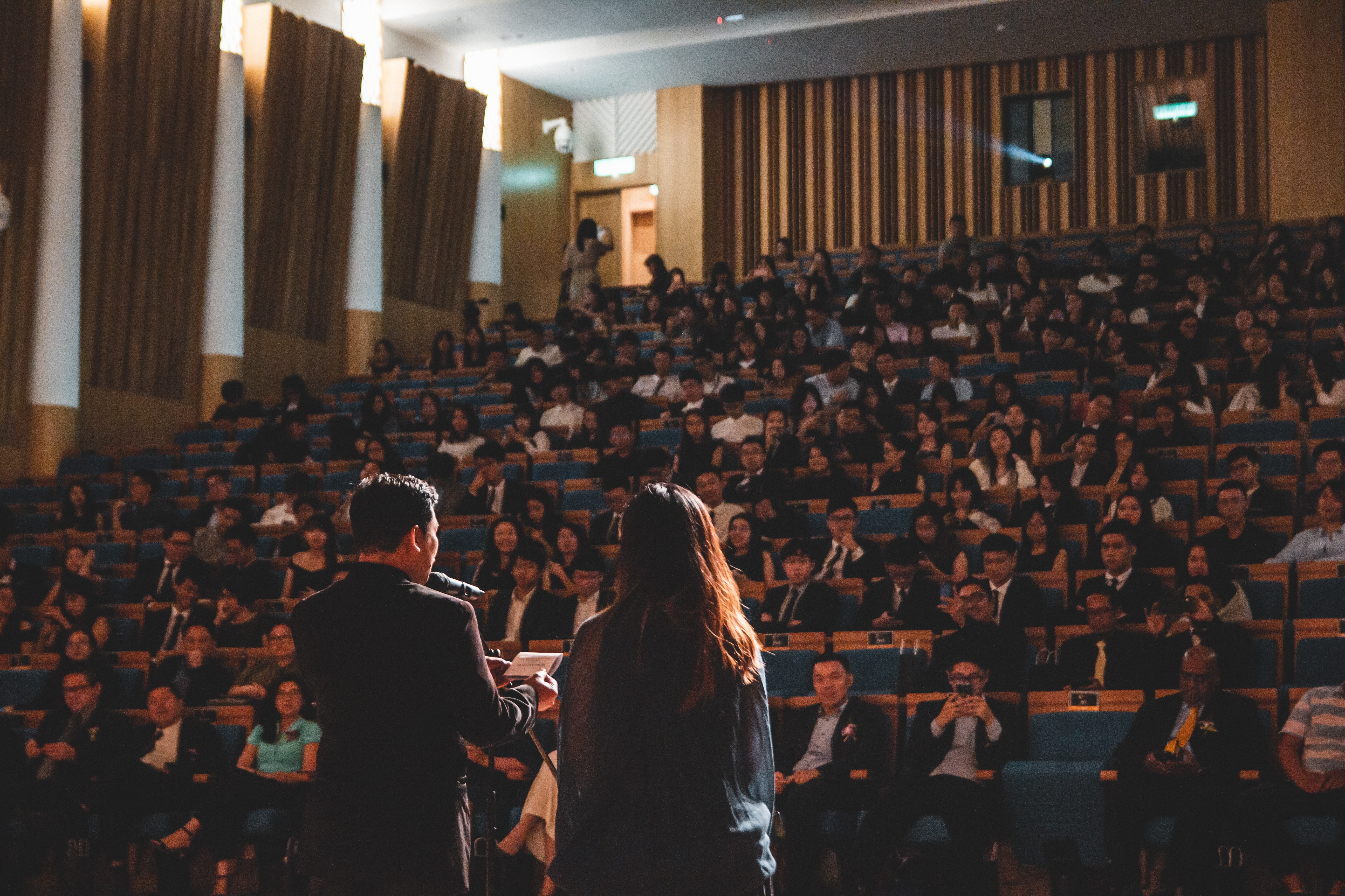 Two people are on stage speaking to a lecture hall that is completely full of hundreds of people