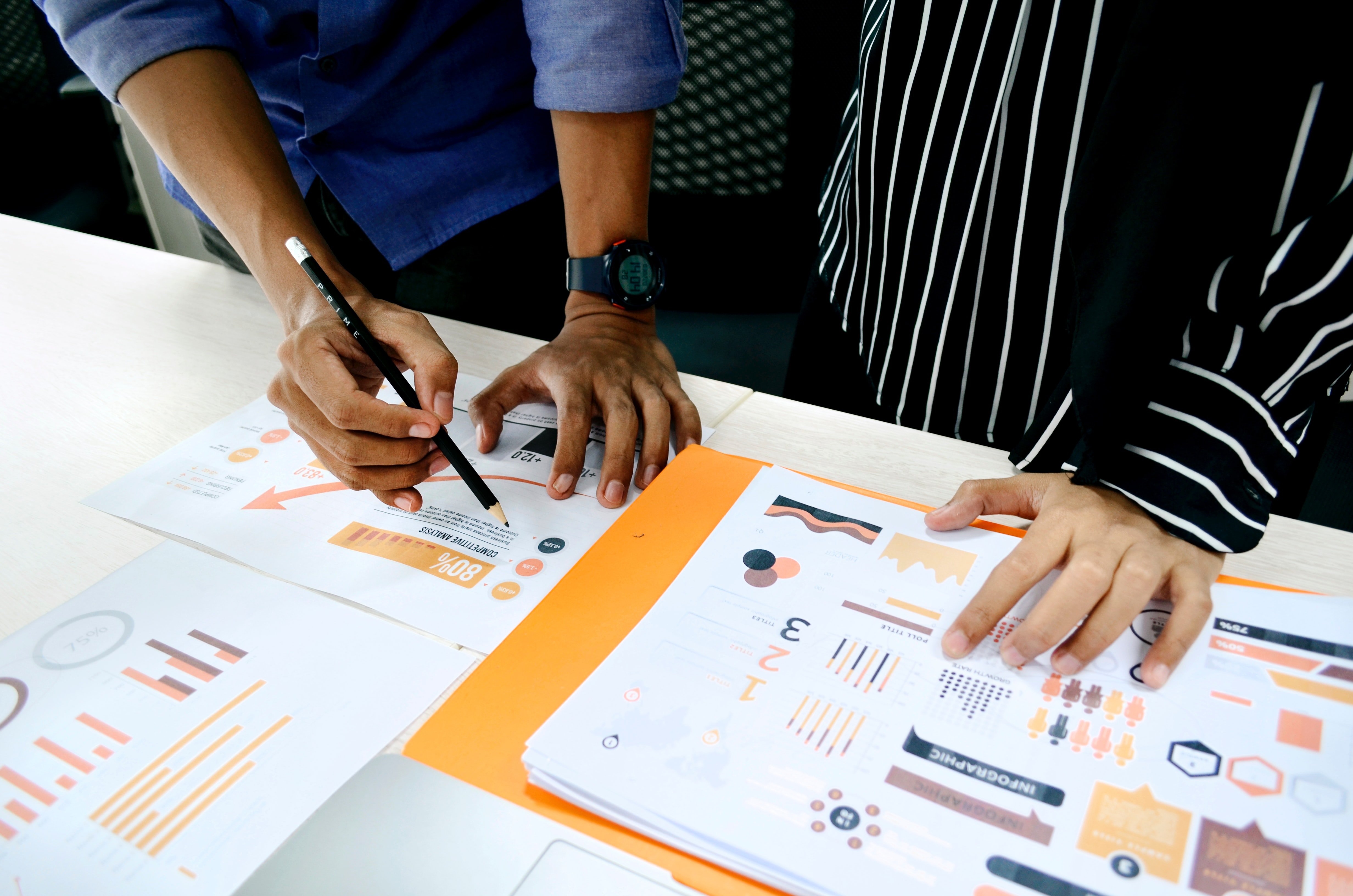 Two people analyze printed data spread out on a desktop and highlight parts with a pencil