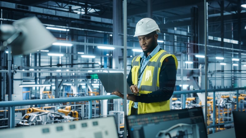 Man wearing hard hat holding laptop in large production facility