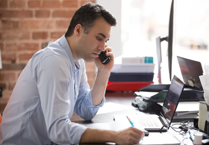 A busy office worker on the phone writing on paper in front of a laptop