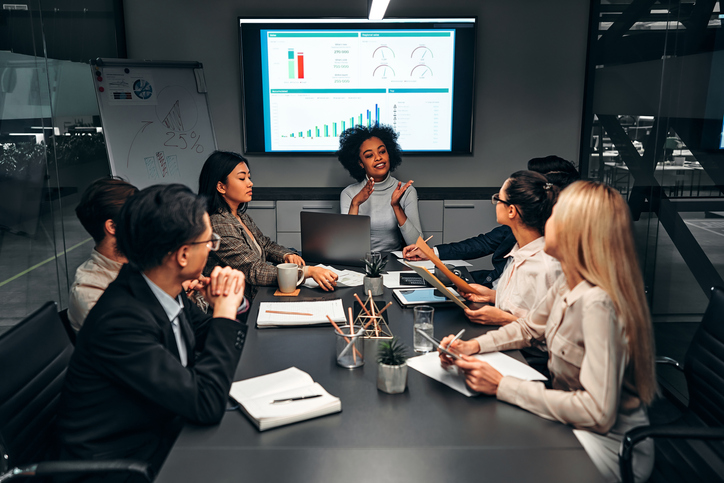 A woman presenting around a conference room with dashboards on a screen