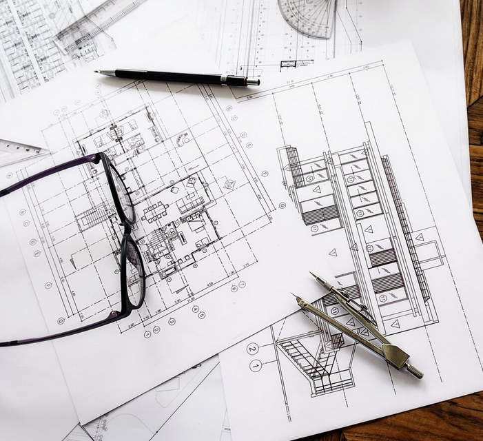 Architectural plans on a table with a compass, protractor, reading glasses, a pen and a ruler on top of them.