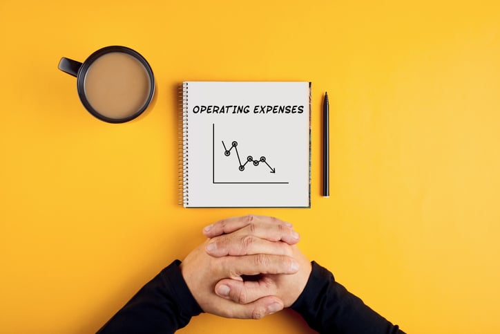 Image of a notebook showing decreasing operating expenses, with a coffee mug and a man's arms also showing