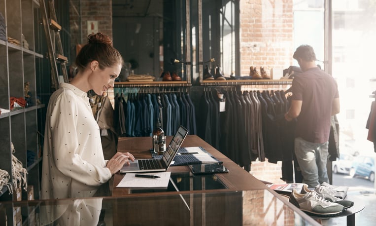 A woman uses a laptop in a clothing store, while a customer browses.