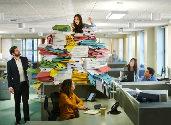 A worker in an office has a large amount of papers on her desk, while others work on their computers.