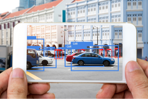 Picture of an object detection application showing cars on a street. The application is running on a phone held by hands in front of the street scene.